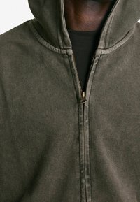 Sudadera con capucha color verde oliva con cierre de cremallera, fabricada con tela texturizada, que cuenta con cierre frontal de cremallera y capucha con cordón. Detalles incluyen costuras visibles.