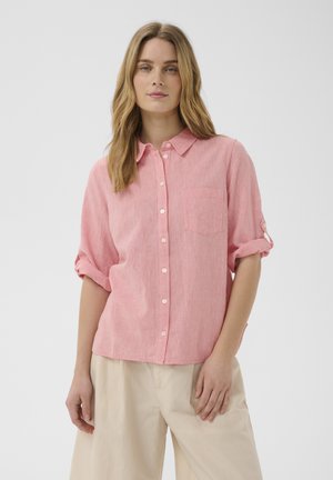 Žena oblečená v svetloružovej košeli na gombíky s vyhrnutými rukávmi a béžových nohaviciach, stojaca pred jednoliatym svetlým pozadím.