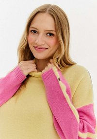 Pletený oversized sveter s žltým telom a ružovými rukávmi. Má vysoký golier a kontrastný pruhovaný vzor pozdĺž rukávov.