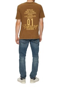 Bruine katoenen t-shirt met grote gele drukletters, met stadsnamen en een grafisch element, gecombineerd met blauwe denim jeans en witte sneakers.