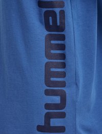 Blå bomullströja med en framträdande svart vertikal logotyp längs vänstra sidan, slät textur och avslappnad design.