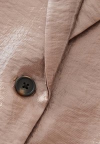 Blush Satin-Stoff mit einem subtilen Glanz, ausgestattet mit einem schwarzen Vierlochknopf und einem taillierten Revers, das eine glatte Textur und Faltendetails zeigt.
