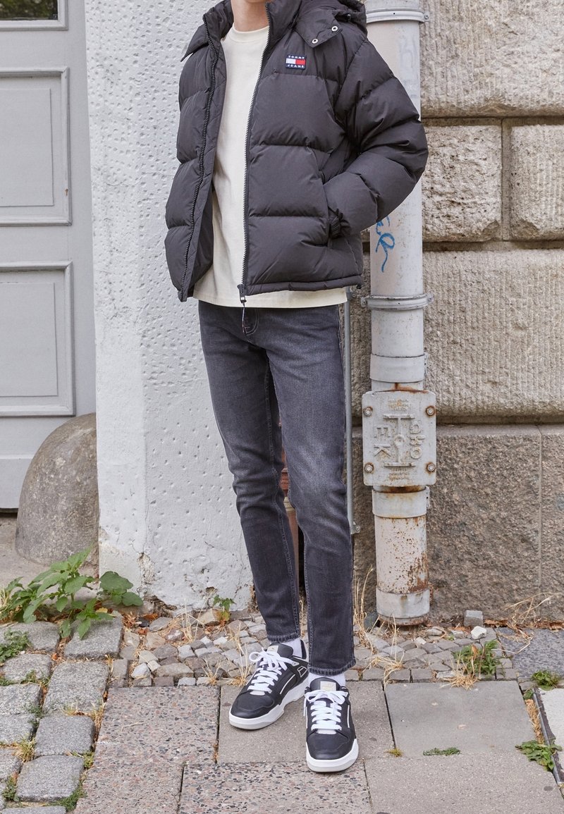 Schwarze Pufferjacke mit Kapuze, darunter ein cremefarbenes Hemd, dunkle Jeans und schwarze Sneaker. Die Jacke hat ein Logo-Patch auf der linken Brust.