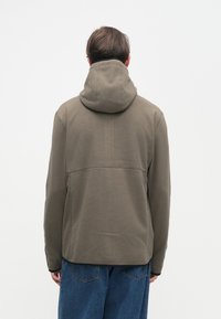 Χακί μπουφάν με κουκούλα, κατασκευασμένο από υφασμάτινη ύλη με υφή και μονόχρωμο πίσω μέρος. Διαθέτει άνετη εφαρμογή και συνδυάζεται με φαρδιά μπλε τζιν.