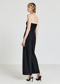 Schwarzer trägerloser Jumpsuit mit tailliertem Oberteil, weitem Hosenbein und subtilen vertikalen Nadelstreifen. Getragen mit High Heel Sandalen.