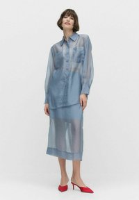 Ljust blå genomskinlig blus med knäppning fram och ficka, parat med en matchande genomskinlig kjol, bärs med röda stilettklackar.