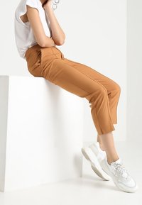 Tan tapered trousers with front pleats, paired with a white short-sleeve top and chunky white sneakers. Smooth fabric, minimalist design.