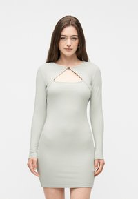 Γυναίκα που φοράει μια μίνι φόρεμα μακρυμάνικο με ριμπ ύφασμα σε ανοιχτό πράσινο χρώμα, με άνοιγμα στο στήθος και λεπτομέρεια με κόπιτσα, στέκεται μπροστά σε ένα απλό φόντο.