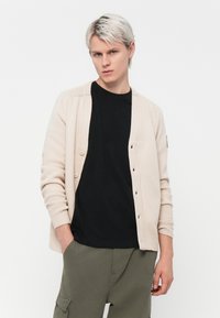 Light beige cardigan with a ribbed texture, button closure, and two front pockets, layered over a black t-shirt and olive green pants.