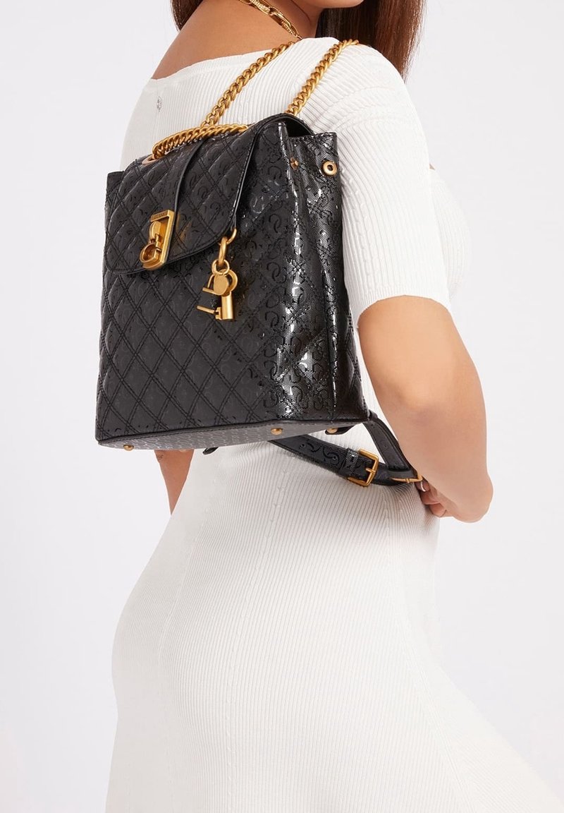 Femme portant une robe blanche, elle porte un sac à dos en cuir noir matelassé avec des bretelles en chaîne dorée et un pendentif cadenas sur une épaule.