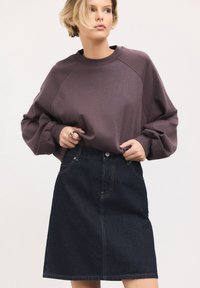 Donna che indossa una felpa viola larga infilata in una gonna a trapezio in denim scuro, in piedi contro uno sfondo chiaro uniforme.