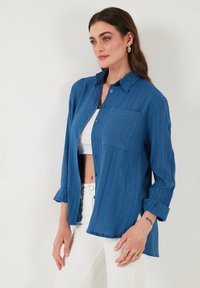 Blau strukturierte Hemdbluse mit einer Brusttasche, langen Ärmeln, die an den Manschetten aufgerollt sind, kombiniert mit einem weißen Cropped-Top und weißen Hosen.