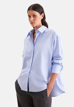 Vrouw met donker haar die een lichtblauw oversized overhemd met knopen en donkergrijze broek draagt, staand met één hand in de zak.