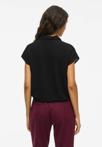 Črn zgornji del s kratkimi rokavi in ovratnikom, teksturirana tkanina, spuščena, v kombinaciji z maroonskimi hlačami. Podrobnosti o malce zavihanih rokavih.