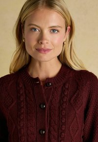 Burgundská pletená vesta s okrúhlym výstrihom, textúrovanými káblovými vzormi a čiernymi gombíkovými uzávermi vpredu. Mäkká, stredne ťažká tkanina.