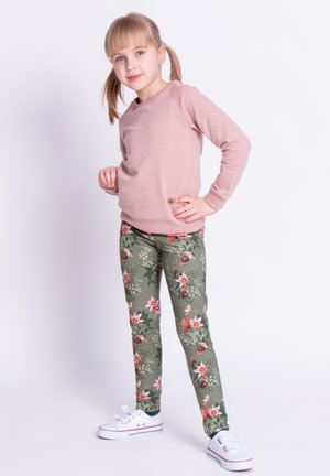 Jasnoróżowa bluza z rękawami raglanowymi i detalem z logo, zestawiona z zielonymi legginsami w kwiaty z różowymi i białymi kwiatami oraz białymi trampkami.