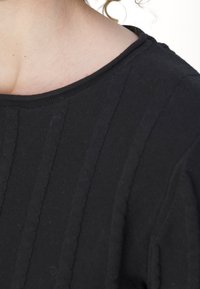 Gros plan sur le cou et l'épaule d’une personne portant un pull côtelé noir avec des mèches de cheveux bouclés lâches visibles.