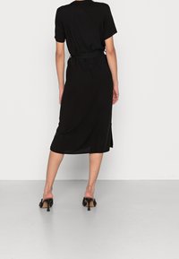 Schwarzes Midikleid mit kurzen Ärmeln und einem taillierten Design. Das Kleid hat eine glatte Textur und seitliche Schlitze, kombiniert mit strappy High-Heel-Mules.