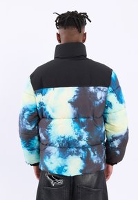 Polstret jakke med en sort topsektion og et multicolor tie-dye mønster i blå og grøn. Har en høj krave og elastiske manchetter.