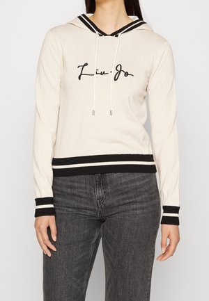 Felpa con cappuccio color crema con polsini e orlo a righe nere, testo in corsivo nero sul davanti, indossata con jeans grigio scuro da una persona in piedi.