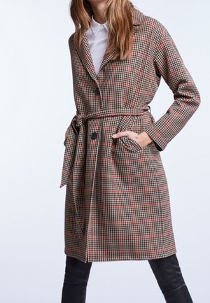 Femme portant un manteau ceinturé à carreaux marron et rouge jusqu'aux genoux avec des revers et des boutons noirs, assorti à un pantalon noir et un haut blanc.