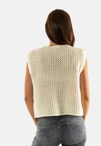 Haut sans manches en maille crochet de couleur blanc cassé, avec un motif ajouré ample et un col arrondi, associé à un jean gris clair.