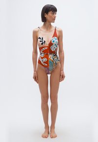 Γυναίκα με κοντά σκούρα μαλλιά που φοράει πολύχρωμο ολόσωμο μαγιό με αφηρημένο σχέδιο και δεμένη ζώνη, όρθια ξυπόλυτη μπροστά σε λευκό φόντο.