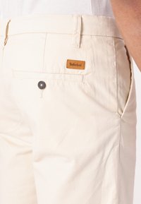 Bež bombažne hlače z zadnjim žepom, imajo črn gumb in svetlo rjavo usnjeno oznako Timberland na pasu. Gladko teksturo.