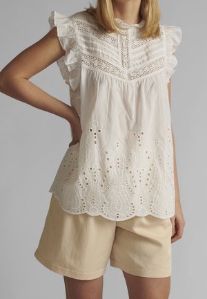Femme portant un chemisier blanc en dentelle sans manches avec des volants et un short beige, debout avec une main sur la hanche sur un fond gris.