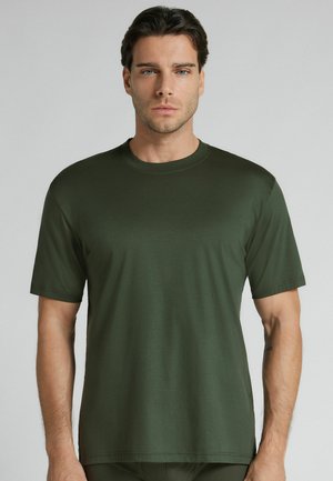 Mand med kort mørkt hår iført en ensfarvet mørkegrøn T-shirt med korte ærmer, stående foran en ensfarvet lys grå baggrund.