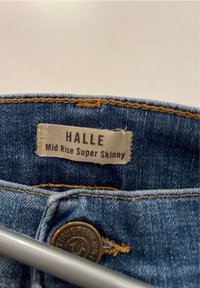 Blue denim jeans with a mid-rise waist, super skinny fit, featuring gold stitching and a branded metal button. Label reads "HALLE."