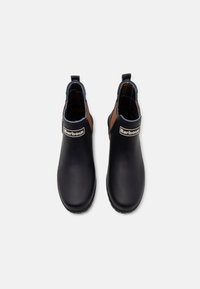 Botas de tornozelo pretas em borracha com uma textura lisa, detalhes castanhos e forro xadrez. Etiqueta do logótipo na lateral e puxador na parte de trás.