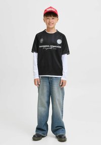 Svart kortärmad tröja med vit text och logotyp, i kombination med en långärmad vit skjorta, vida blå denimjeans och svarta skor. Röd keps på huvudet.