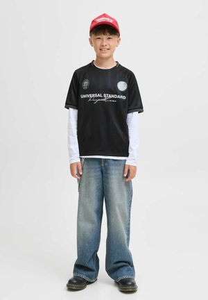 Black short-sleeve jersey with white text and logo, paired with a long-sleeve white shirt, wide blue denim jeans, and black shoes. Red cap on head.
