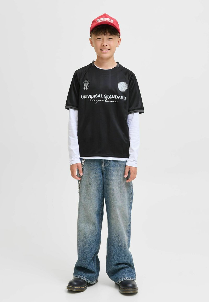 Svart kortärmad tröja med vit text och logotyp, i kombination med en långärmad vit skjorta, vida blå denimjeans och svarta skor. Röd keps på huvudet.