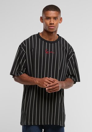 Young man with short hair wearing a black pinstriped oversized T-shirt with red cursive text, standing with hands clasped against gray background.