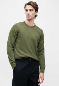 Νεαρός άνδρας που φοράει ένα ελιά-πράσινο μπλουζάκι με στρογγυλή λαιμόκοψη και μαύρο παντελόνι, στέκεται με το ένα χέρι στην τσέπη, κοιτάζοντας ελαφρώς προς τα δεξιά.