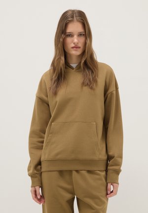 Junge Frau mit glattem hellbraunem Haar, die einen übergroßen braunen Hoodie und passende Hose trägt, steht vor einem einfarbigen Hintergrund.