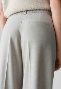 Pantalons ajustés gris clair avec une finition texturée, comportant deux poches arrière et un design à plat à l'avant. Détail de ceinture caché visible.
