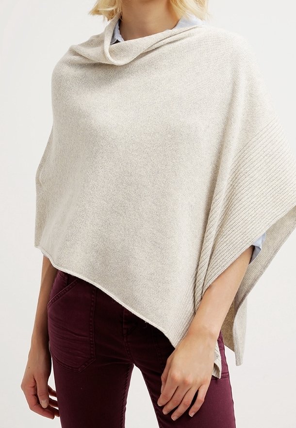 Femme portant un poncho en tricot beige clair par-dessus une chemise, associé à un pantalon ajusté violet foncé.