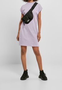 Lawendowa sukienka z krótkim rękawem o luźnym kroju, zestawiona z czarną torebką typu crossbody. Obuwie stanowią czarne botki za kostkę o teksturowanym wykończeniu.