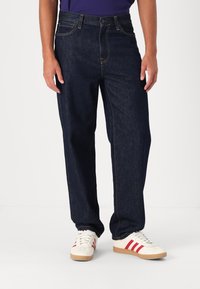Vaqueros de mezclilla azul oscuro con corte recto, costuras visibles y bolsillos delanteros. Combinados con zapatillas blancas con rayas rojas.