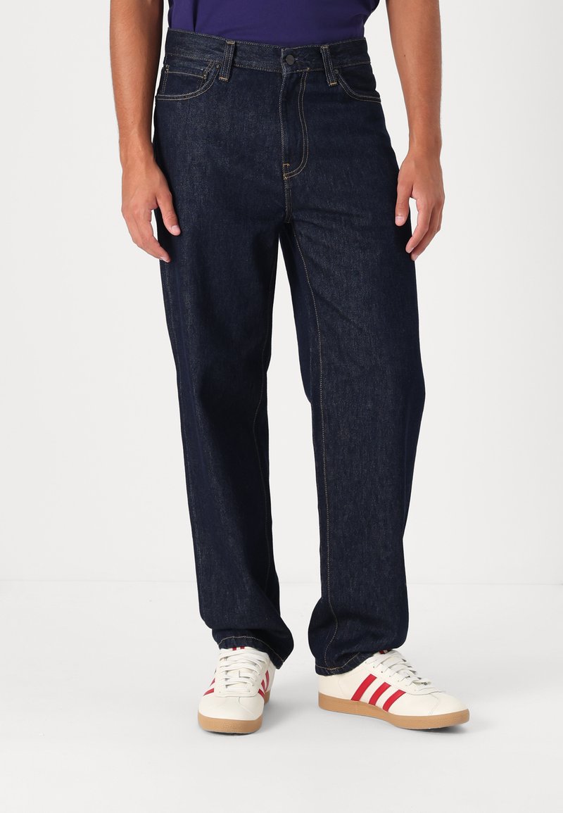 Vaqueros de mezclilla azul oscuro con corte recto, costuras visibles y bolsillos delanteros. Combinados con zapatillas blancas con rayas rojas.