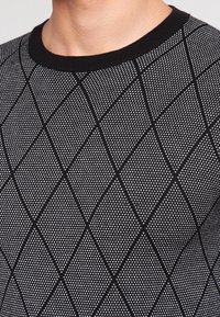 Pull en tricot noir et gris présentant un motif en losange avec de petits motifs triangulaires et un col côtelé noir.