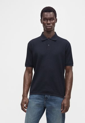 Mandlig model iført en mørk navy kortærmet ribbet poloshirt og blå jeans, stående foran en ensfarvet lys baggrund.