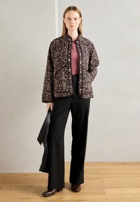 Woman wearing a black floral quilted jacket, mauve ribbed top, black wide-leg trousers, brown shoes, holding a black bag, standing on wooden floor.
