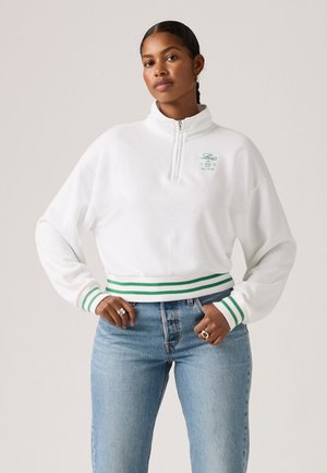 Woman wearing white quarter-zip sweatshirt with green stripes on cuffs and hem, small embroidered logo, and light blue jeans, standing with hands on hips.