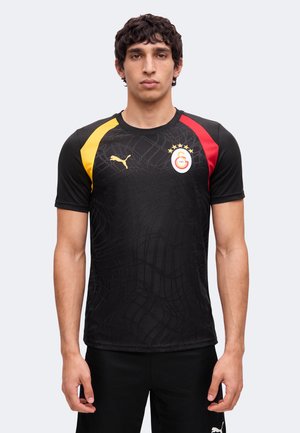 Homme portant un maillot de sport noir Puma avec des bandes jaunes et rouges aux épaules et le logo du club de football Galatasaray sur la poitrine.