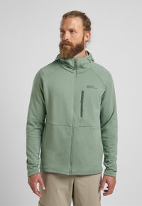 Veste zippée vert clair en tissu texturé, avec une capuche et une poche poitrine. Présente un logo sur le côté gauche et des manches ajustées.