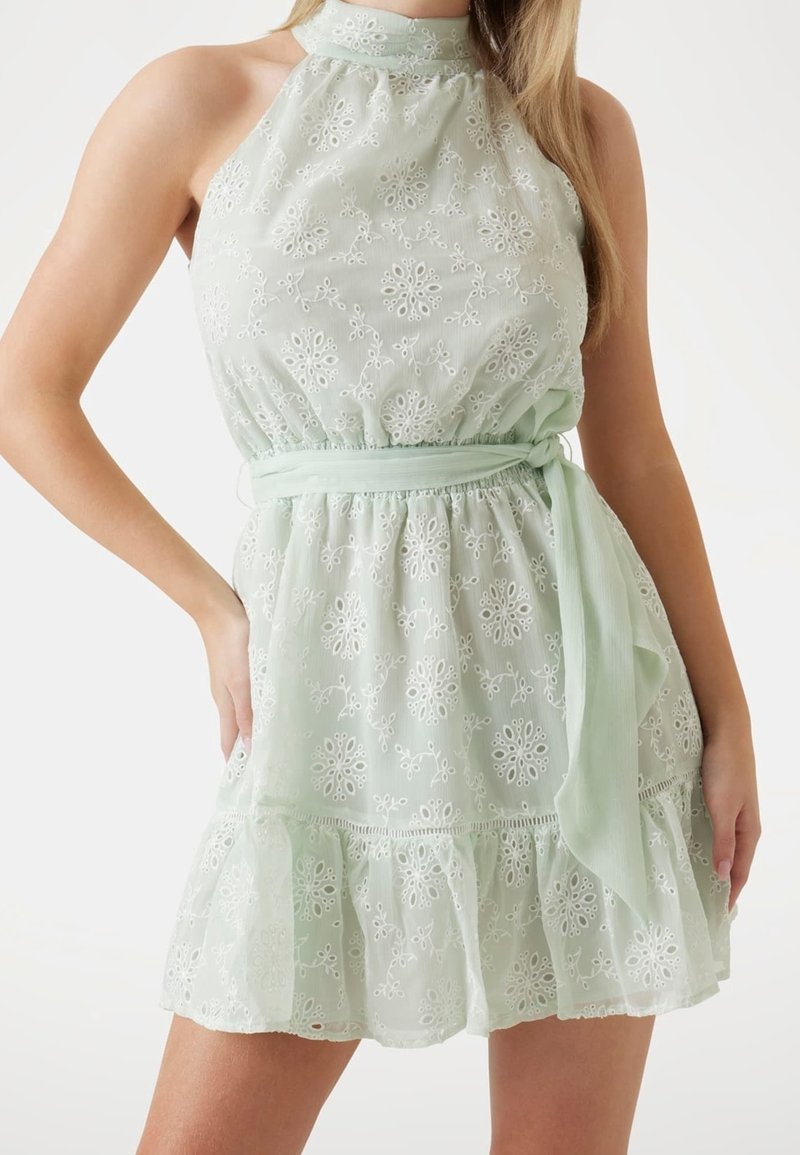 Femme portant une robe en dentelle vert pastel sans manches, col montant, broderies florales, ceinture à la taille et ourlet volanté.